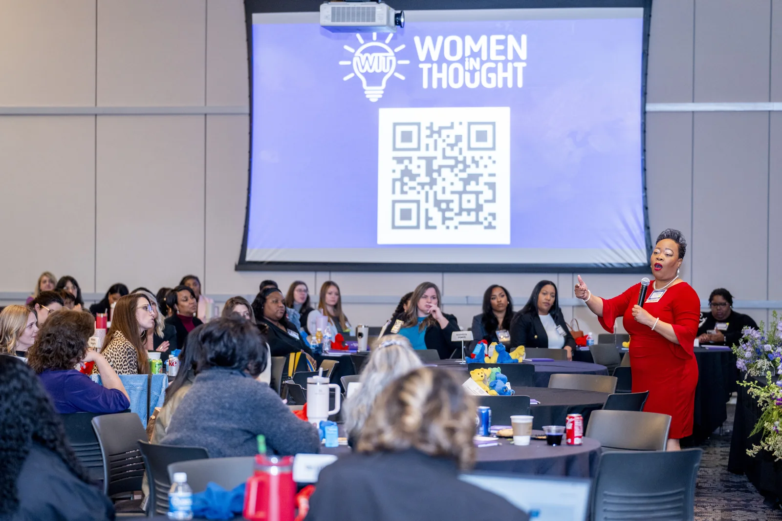 Women in Thought Summit room with attendees, presentation screen, table materials, and branded event environment visible.
